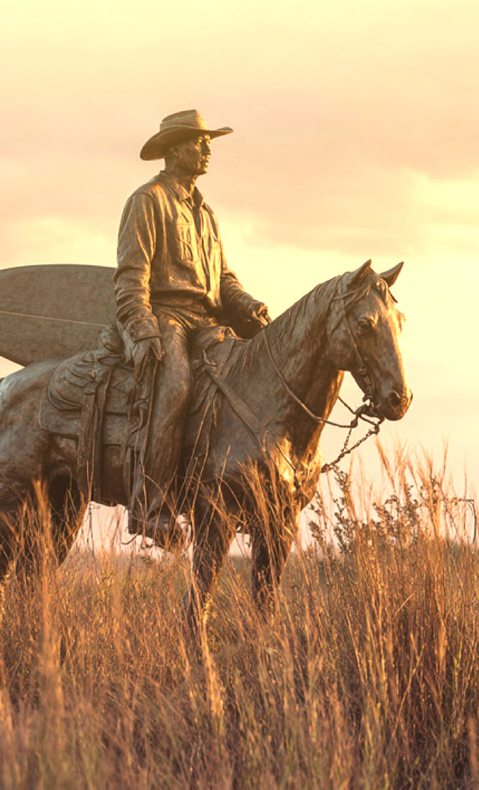 A rider on horseback carrying a surfboard through tall grass at sunset