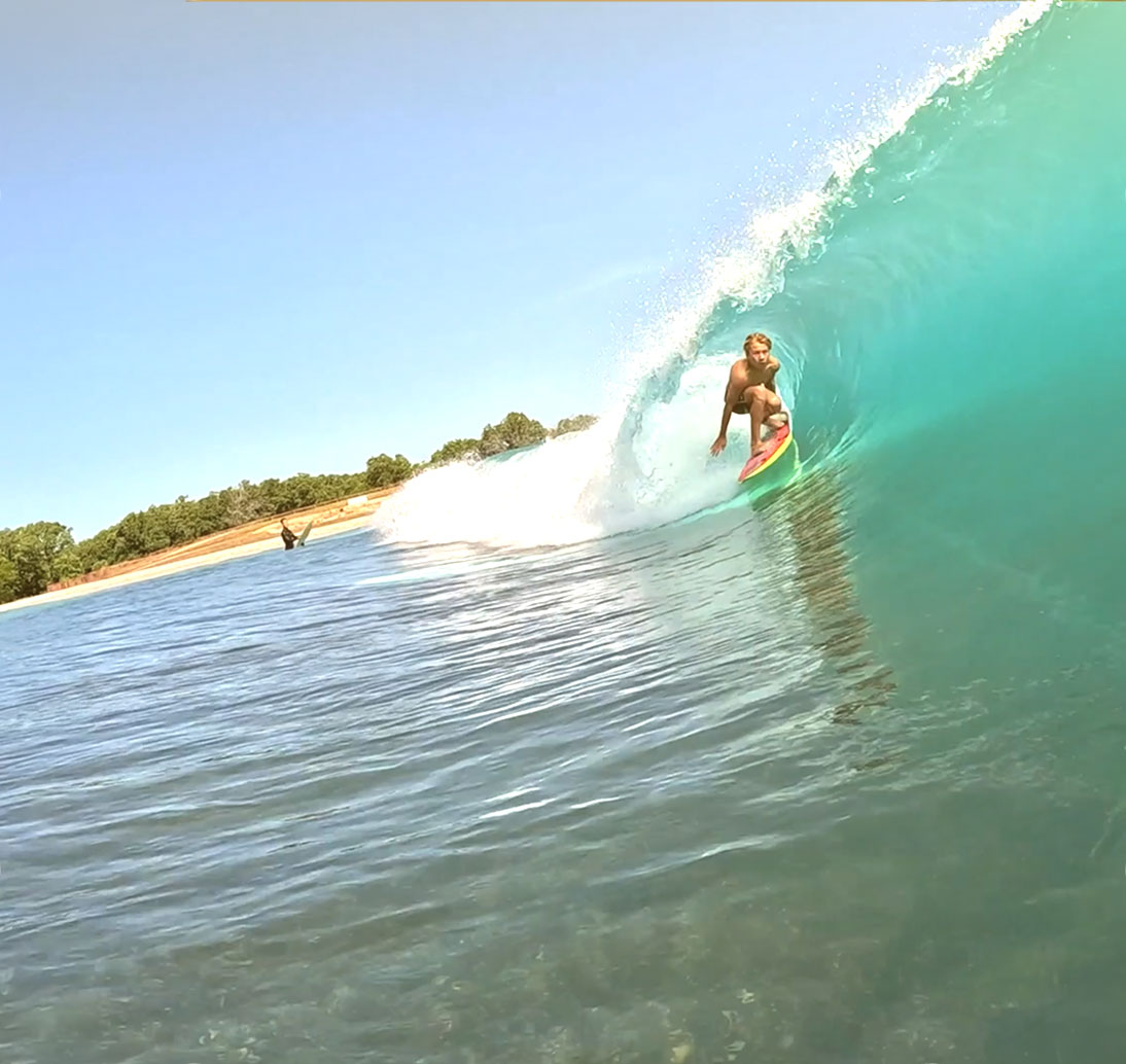 A surfer riding deep in the curl of a head-high wave on the Waco Surf lagoon, the tree-lined bank low behind it